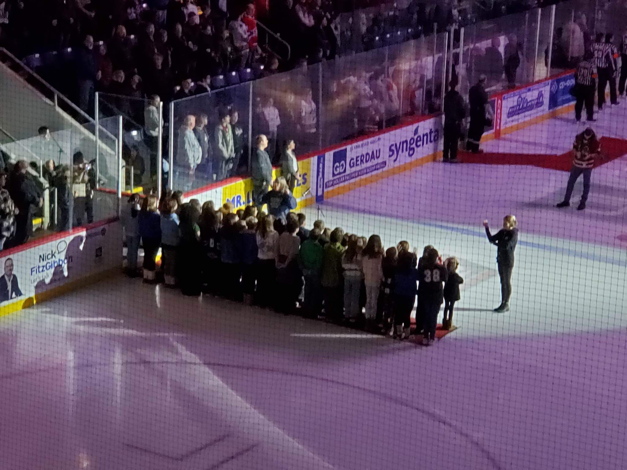 This photo features a group of elementary age students standing on the ice of a hockey rink, with their teacher to the side in front of a large audience.