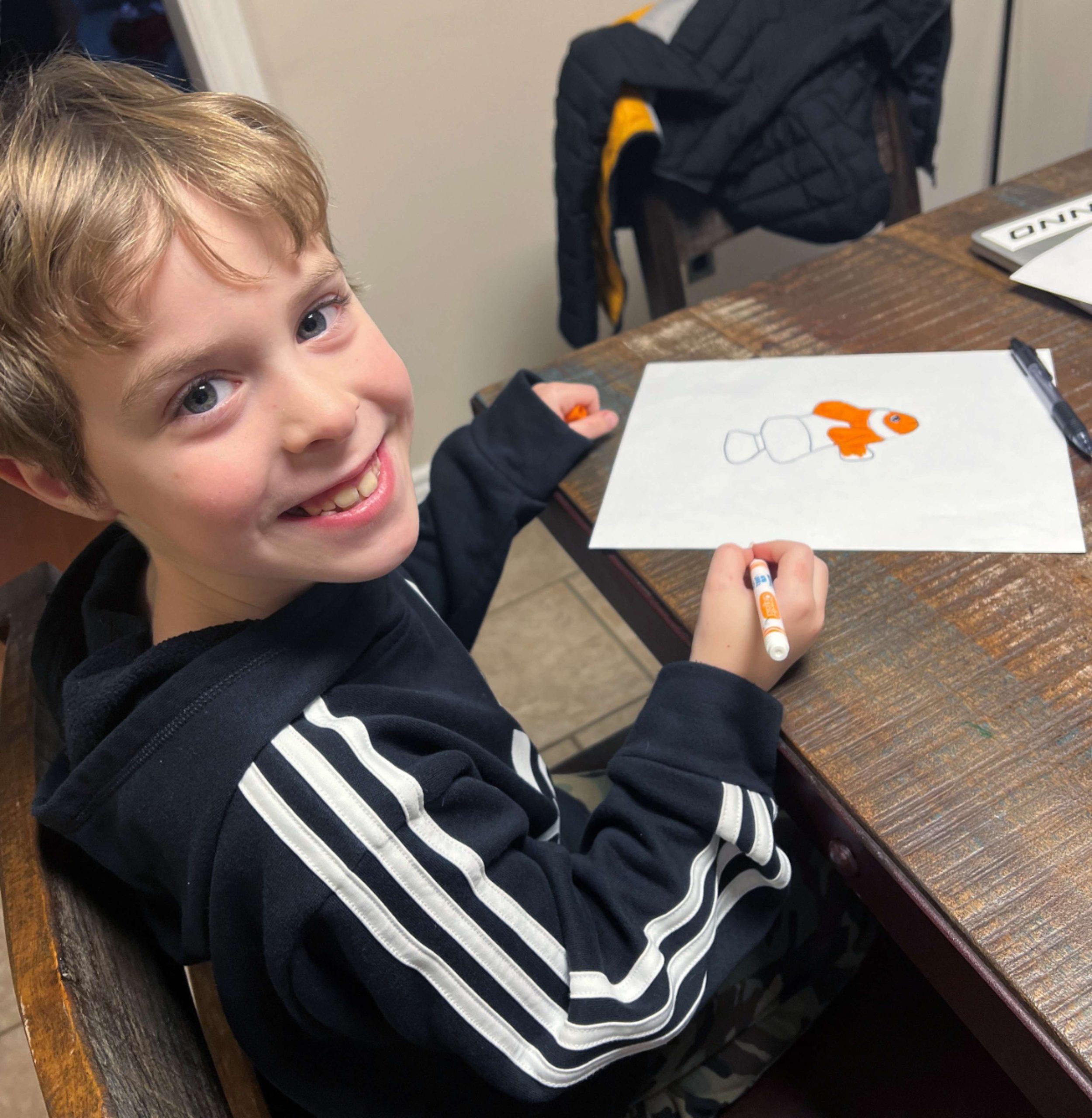 This photo features a young blonde boy smiling at the camera while sitting around a kitchen table where he works on a hand drawing of a clownfish. 
