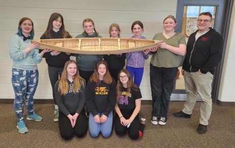 This picture features a group of nine girls alongside their teacher with finished canoe model project