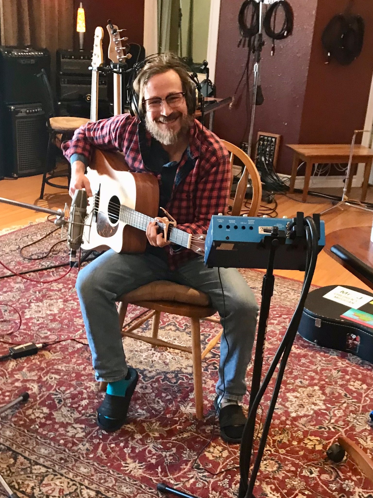 This photo features musician Geoff Jackson sitting in the middle of a recording room holding an acoustic guitar. 