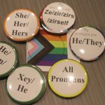 This photo shows 6 buttons with different pronouns around a small Progressive Pride Flag.