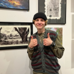 This photo shows Neilan C standing in front of their artwork with their thumbs up.