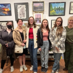 This photo features a group of students and their teacher smiling in front of pictures of art.