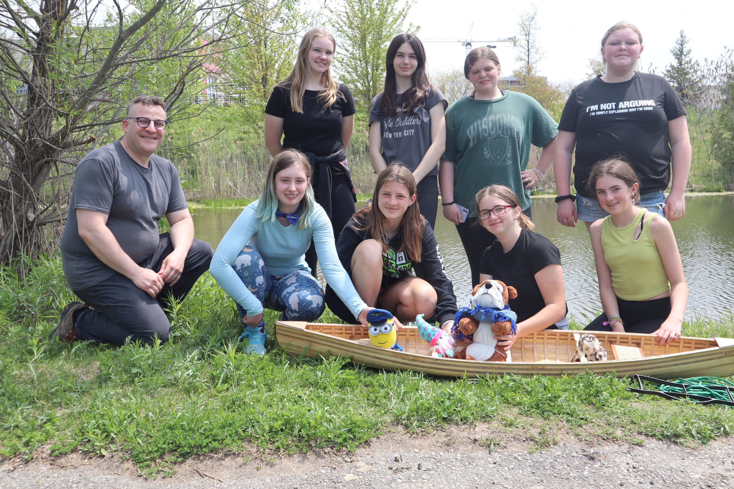 This photo features a group of 8 female students and 1 male teacher smiling and posing with their model canoe filled with stuff toys.