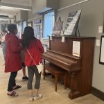 This photo features 2 students looking at artwork behind a piano.