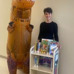 This photo features a woman in a dinosaur suit next to another women holding a mobile cart with books on it and a caption that reads "TJRC letting staff know that school libraries are not extinct."